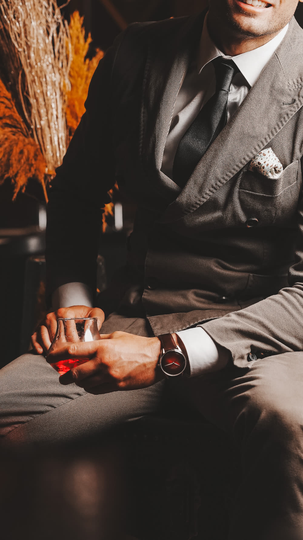 A man in a tailored grey suit wearing an ELKA watch on a brown leather strap, holding a glass in a candlelit bar